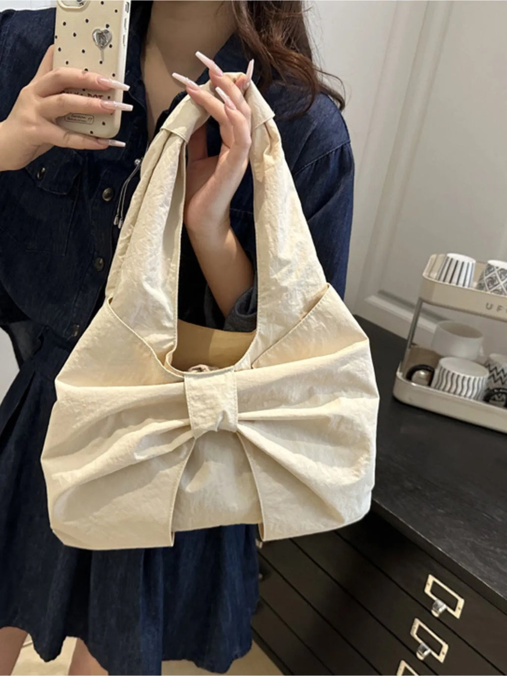Cream bow shoulder bag held by its strap in a front mirror view.