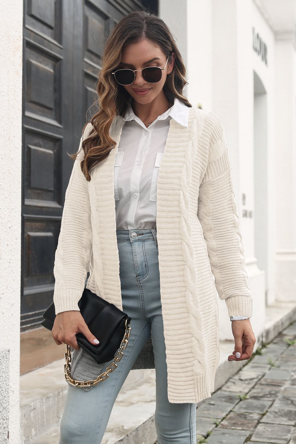 Ivory long open-front cable-knit cardigan worn with a white shirt, jeans and a black chain-strap bag.