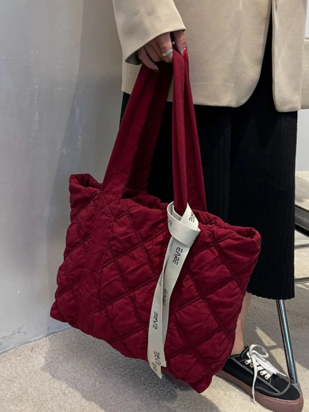 Burgundy quilted tote held by its handles beside the model's legs.