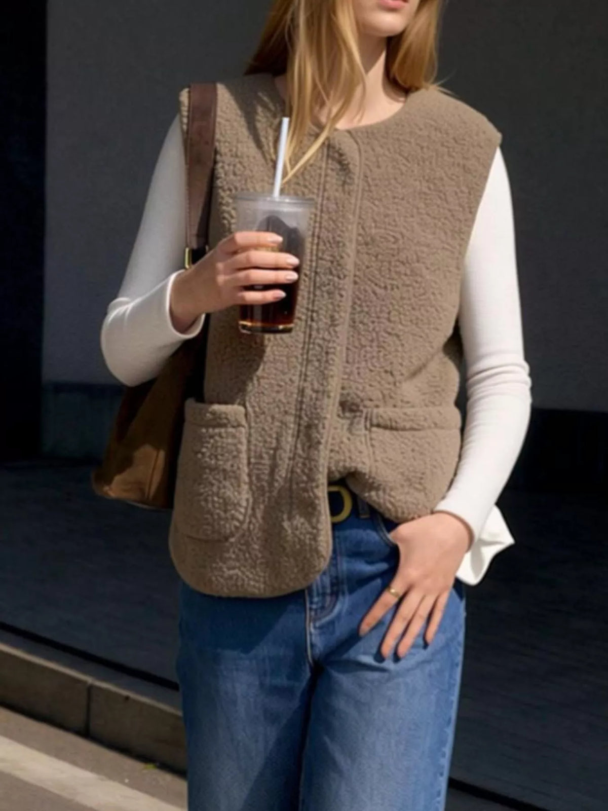 Khaki sherpa vest worn open over a white top with jeans, holding an iced coffee.