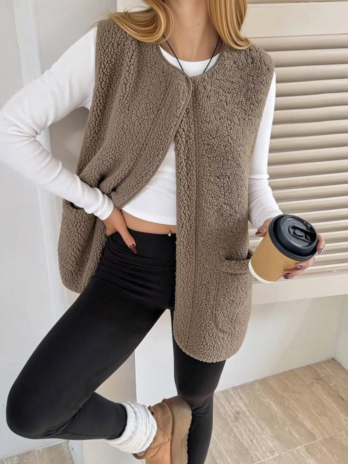 Khaki sherpa vest worn open with black leggings, holding a coffee cup.